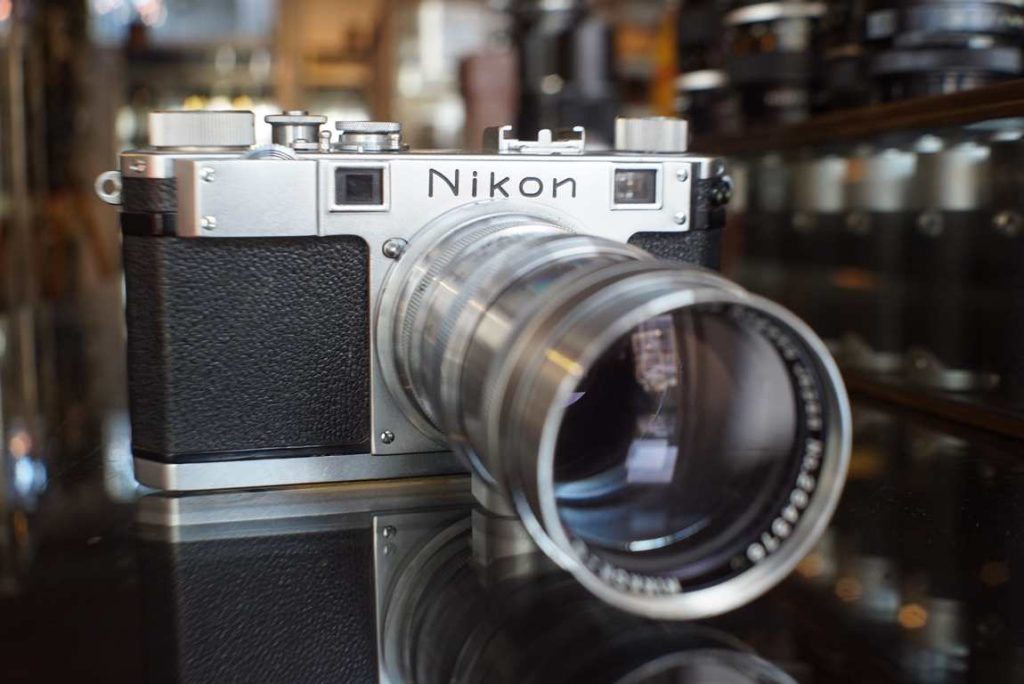 Nikon S rangefinder camera with matching telephoto 135mm F/3.5 lens. Sold for collection or restoration purposes. The lens is a Nippon Kogaku Nikkor-Q.C f=135mm 1:3.5 version. The lens does have a light layer of dust inside but this could easily be cleaned if opened. The camera has perfect cosmetics and some normal age related scuffs there and there, overall in a very well preserved and good looking condition. The shutter does fire but is very sluggish and irregular, there are some wrinkles in the curtain. It will not expose film correctly and would benefit from a shutter service and probably full CLA. Front plate of camera looks very clean, no marks there, clean rangefinder windows, no issues there at all. Topplate shows minimal wear and buttons feel responsive. Focus wheel is very heavy though, but the focus ring on the lens works beautiful. Sold without warranty.