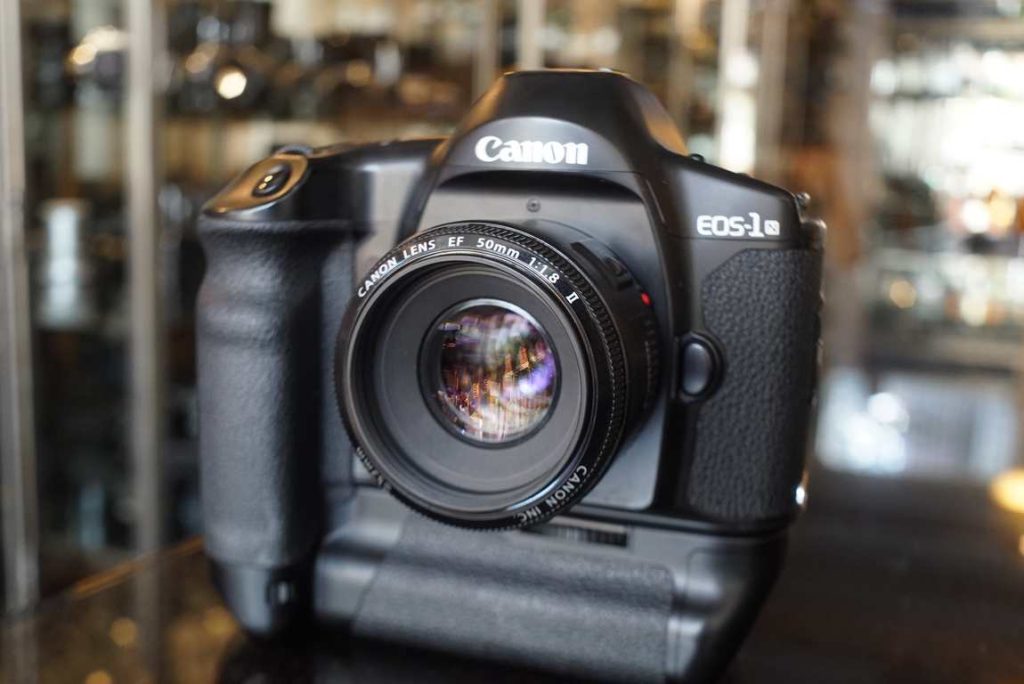 Canon EOS 1N with PB-E1, or also called the EOS 1N HS, comes with a EF 50mm f/1.8 II. The 1N has 5 autofocus points and can shoot up to 6 fps with the attached PB-E1 The body in excellent condition with just light marks of use. The camera was machine tested and all speeds operate in margin and the lightmeter is accurate. The eyecup is missing but can still be easily found online. The 50mm f/1.8 II is in excellent condition with minimal wear on the body. Optics look good without fungus or haze. A nice kit for the user as you are able to use all your modern EF glass with this camera.