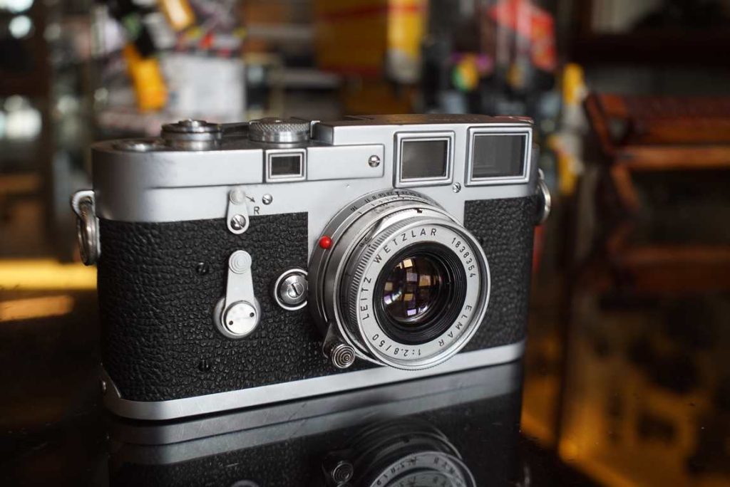 This Leica M3 dual stroke camera has been produced in 1955 and comes with an Elmar 50mm f/2.8 M lens from 1961. The camera comes in good cosmetical condition for it's age. Regular signs from previous use. Some slight meter wear on the top plate and there is 1 bit of vulcanite missing on the back near one of the strap lugs. The transport feels precise and the self-timer also works perfectly. The shutter speeds were checked on our test equipment and speeds up to 1/250 are within tolerance. faster speeds are uneven. The rangefinder image is also a bit dim. The camera is usable as-is if you stay under 1/250, but will need a service if you want to fully enjoy this camera and hence is priced accordingly. The elmar lens is in good condition with light coating marks on the front element but no haze. Focus turns smoothly and aperture ring has full stop clicks too.
